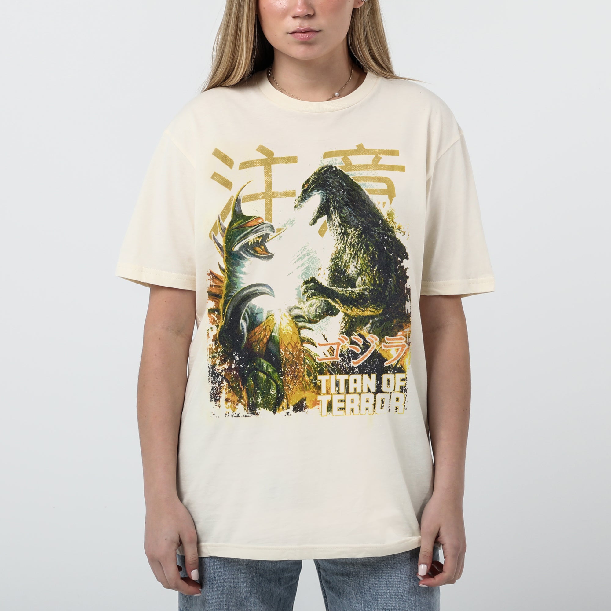 Female modeling Natural-colored T-shirt featuring a vintage-style graphic of Godzilla and Gigan in battle, with Japanese text and "Titan of Terror" lettering.